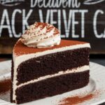 "A rich slice of decadent Black Velvet Cake with smooth cream cheese frosting on a white plate."