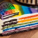 "A vibrant Rainbow Oreo Cheesecake Cake with colorful layers, topped with whipped cream and sprinkles, set on a white plate."