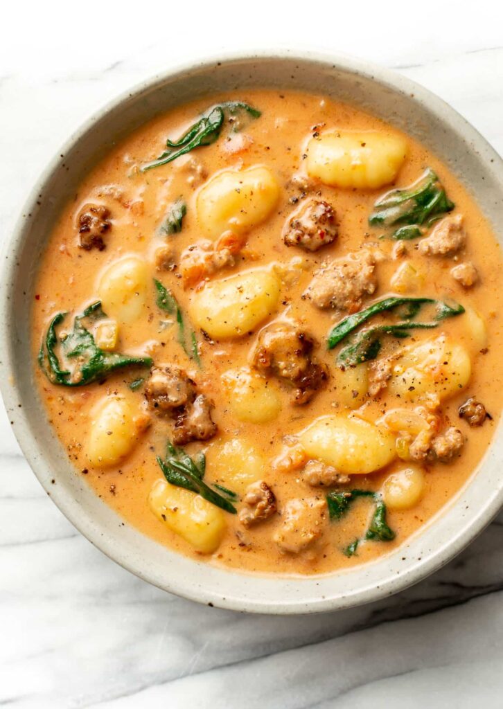Ultra-realistic creamy gnocchi soup with Italian sausage slices, gnocchi, spinach, onions, and herbs in a light creamy broth, served in a ceramic bowl, studio lighting, neutral background